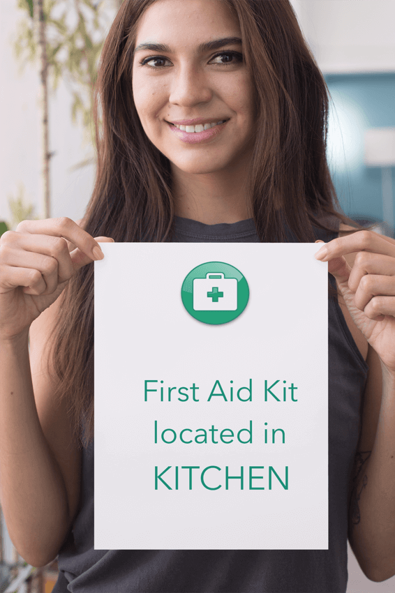 Person holding up sign that says the first aid kit is located in the kitchen